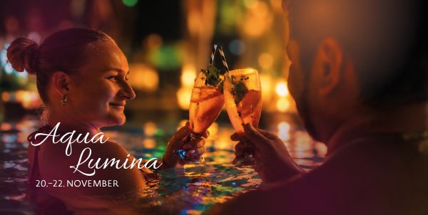 Ein Paar stößt mit Cocktails in der Lagune an, im Hintegrund Kerzenschein. "Aqua Lumina 20. bis 22. November"